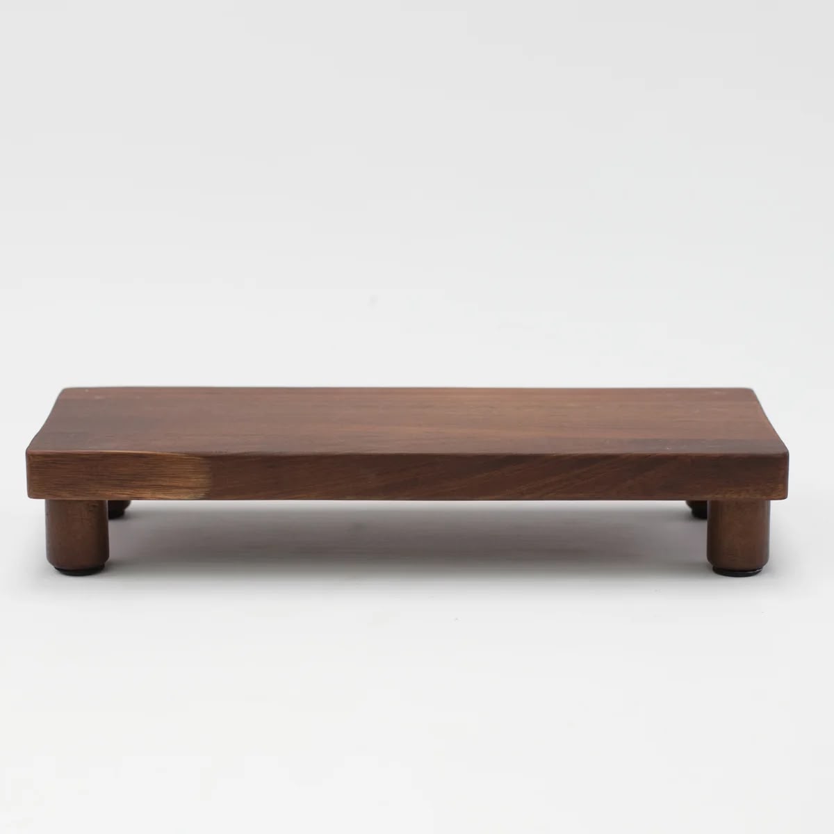 A low walnut riser with turned feet, lifting the back row of a shelf